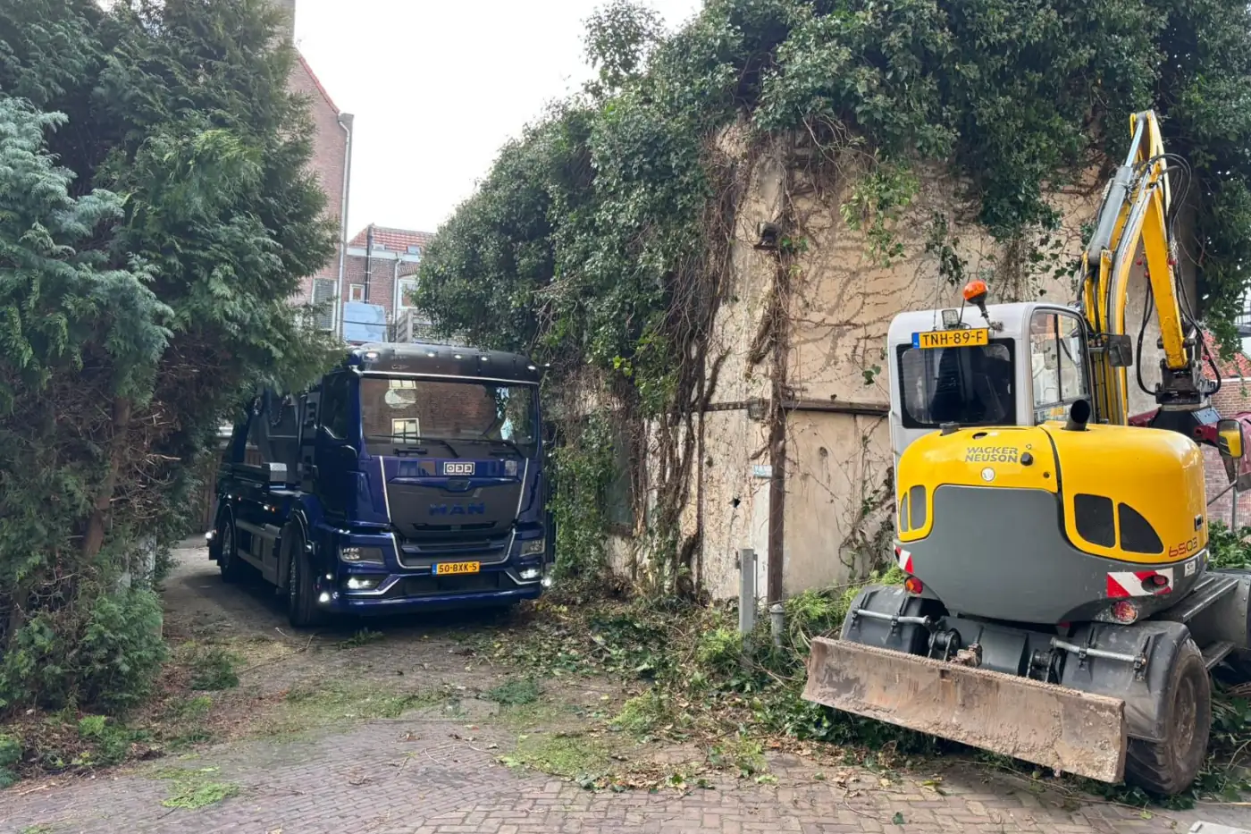 Blauwe 033 Containers containerwagen en gele Wacker Neuson minigraver bij sloop- en grondwerk in een smalle straat