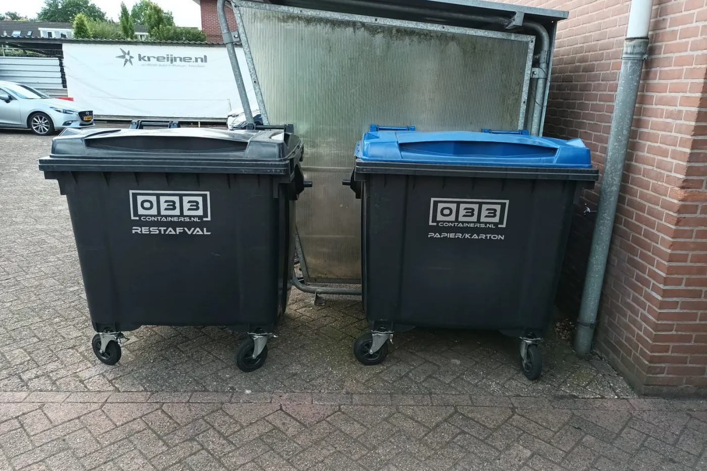 Twee 033 Containers rolcontainers tegen een muur: een zwarte container voor restafval en een container met blauwe deksel voor papier en karton