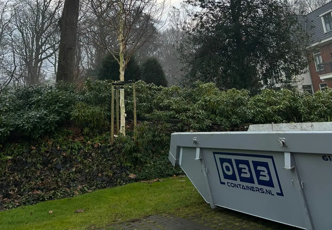 Grijze grondafval container van 033 Containers geplaatst op het gazon bij een tuin met struiken en bomen